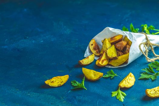 Baked potato wedges on paper with addition sea salt and parsley on a dark blue concrete background