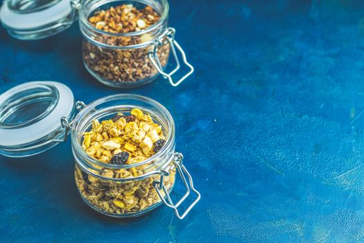 Two open glass jars of organic granola with berries, coconut chips and seeds on a dark blue concrete table surface.