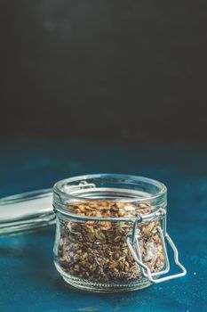 Open glass jar of organic granola with berries, coconut chips and seeds on a dark blue concrete table surface.