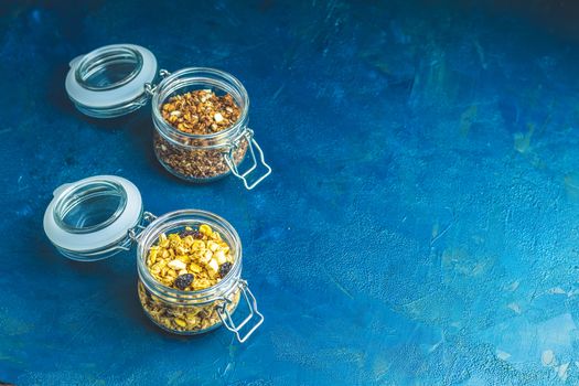 Two open glass jars of organic granola with berries, coconut chips and seeds on a dark blue concrete table surface.