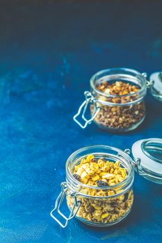 Two open glass jars of organic granola with berries, coconut chips and seeds on a dark blue concrete table surface.