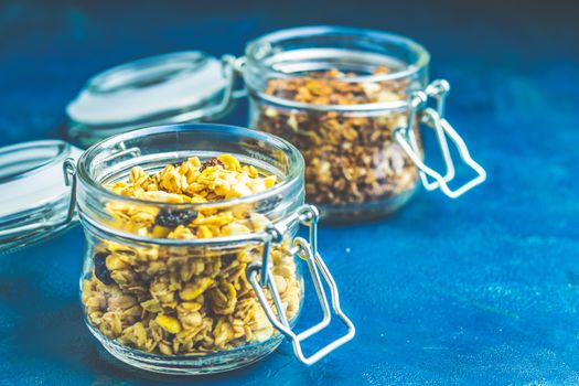 Two open glass jars of organic granola with berries, coconut chips and seeds on a dark blue concrete table surface.