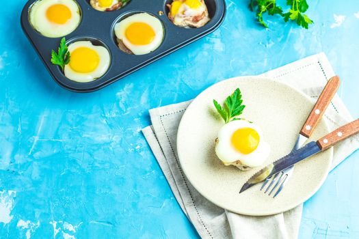 Baked eggs  in light plate and baking molds. Portioned casserole from bacon  and eggs in Italian style. Blue concrete table surface background.