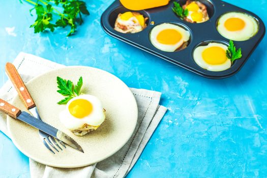 Baked eggs  in light plate and baking molds. Portioned casserole from bacon  and eggs in Italian style. Blue concrete table surface background.