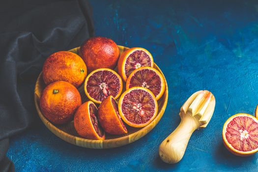 Sliced and whole Sicilian Blood oranges fruits in wooden plate and juicer over dark blue concrete table surface. Dark rustic style.