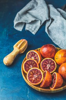 Sliced and whole Sicilian Blood oranges fruits in wooden plate and juicer over dark blue concrete table surface. Dark rustic style.