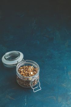 Open glass jar of organic granola with berries, coconut chips and seeds on a dark blue concrete table surface.