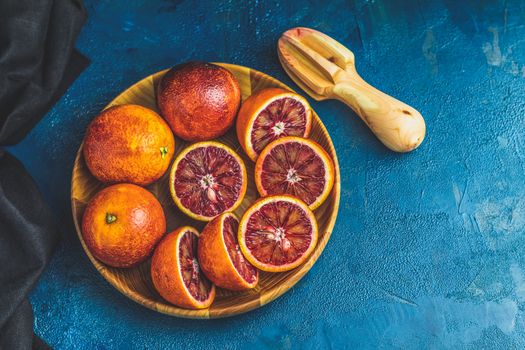 Sliced and whole Sicilian Blood oranges fruits in wooden plate and juicer over dark blue concrete table surface. Dark rustic style.