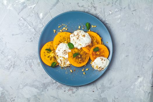 Delicious healthy fruit breakfast. Sliced persimmon with yogurt, brown sugar, pine nuts and fresh mint in blue plate on light gray concrete table surface background, top view, flat lay.