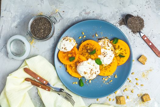 Delicious healthy fruit breakfast. Sliced persimmon with yogurt, chia seeds, brown sugar, pine nuts and fresh mint in blue plate on light gray concrete table surface background, top view, flat lay.