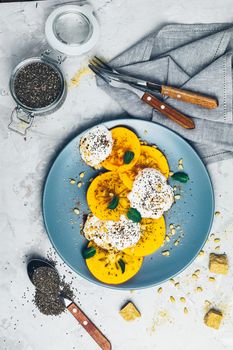 Delicious healthy fruit breakfast. Sliced persimmon with yogurt, chia seeds, brown sugar, pine nuts and fresh mint in blue plate on light gray concrete table surface background, top view, flat lay.