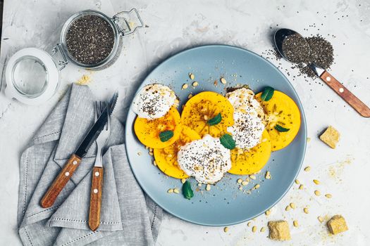 Delicious healthy fruit breakfast. Sliced persimmon with yogurt, chia seeds, brown sugar, pine nuts and fresh mint in blue plate on light gray concrete table surface background, top view, flat lay.