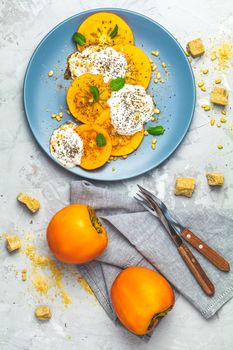 Delicious healthy fruit breakfast. Sliced persimmon with yogurt, chia seeds, brown sugar, pine nuts and fresh mint in blue plate on light gray concrete table surface background, top view, flat lay.