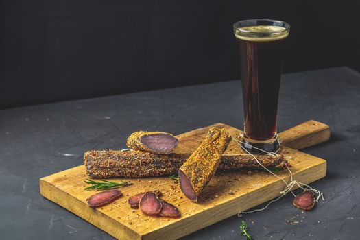 Dark beer in glass and Jerky, basturma, dried meat beef, meat smoked jerky with spices on wooden cutting board, black concrete surface table background.