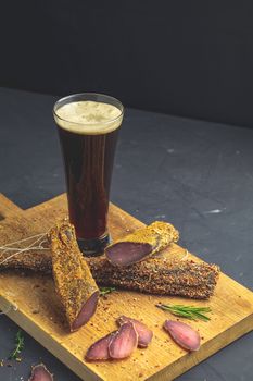 Dark beer in glass and Jerky, basturma, dried meat beef, meat smoked jerky with spices on wooden cutting board, black concrete surface table background.