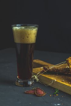 Dark beer in glass and Jerky, basturma, dried meat beef, meat smoked jerky with spices on wooden cutting board, black concrete surface table background.