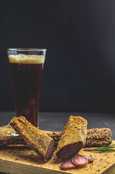 Dark beer in glass and Jerky, basturma, dried meat beef, meat smoked jerky with spices on wooden cutting board, black concrete surface table background.