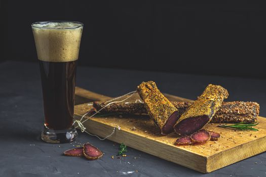 Dark beer in glass and Jerky, basturma, dried meat beef, meat smoked jerky with spices on wooden cutting board, black concrete surface table background.