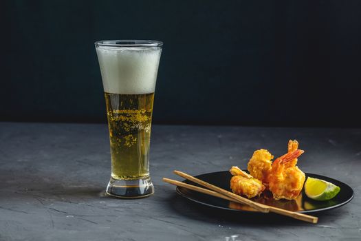 Fried Shrimps tempura with lime in black plate and glass of light beer on dark concrete surface background. Copy space. Seafood tempura dish served japanese or eastern Asia style with chopsticks