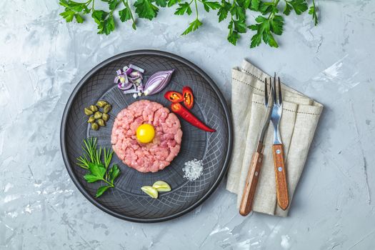 Steak tartare with yolk and ingredients on black ceramic plate, set of cutlery knife and fork on light gray stone concrete textured surface background. Copy space background, top view flat lay.
