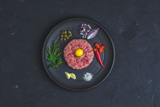 Steak tartare with yolk and ingredients on black ceramic plate, set of cutlery knife and fork on black stone concrete textured surface background. Copy space background, top view flat lay.