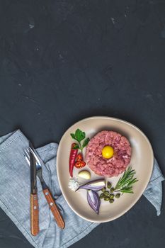 Delicious steak tartare with yolk and ingredients on ceramic plate, set of cutlery knife, fork on black stone concrete textured surface background. Copy space background, top view flat lay.