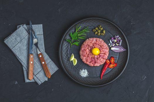 Steak tartare with yolk and ingredients on black ceramic plate, set of cutlery knife and fork on black stone concrete textured surface background. Copy space background, top view flat lay.