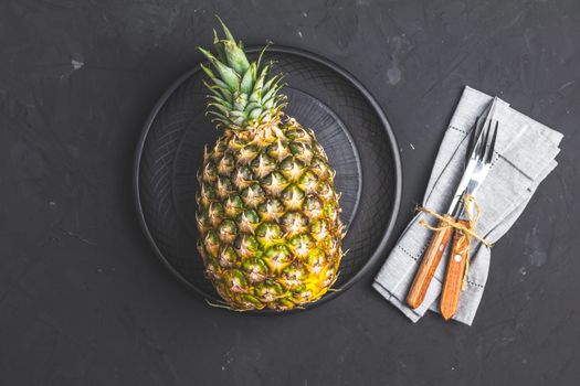 Pineapple in ceramic plate and set of cutlery knife, fork  on black stone concrete textured surface background. Top view with copy space for your text.