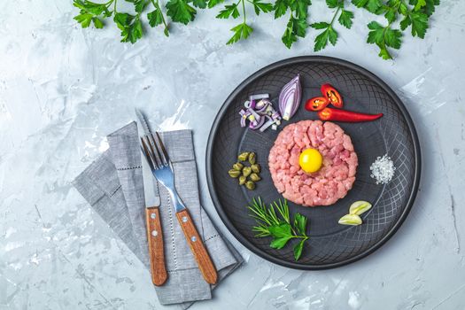 Steak tartare with yolk and ingredients on black ceramic plate, set of cutlery knife and fork on light gray stone concrete textured surface background. Copy space background, top view flat lay.