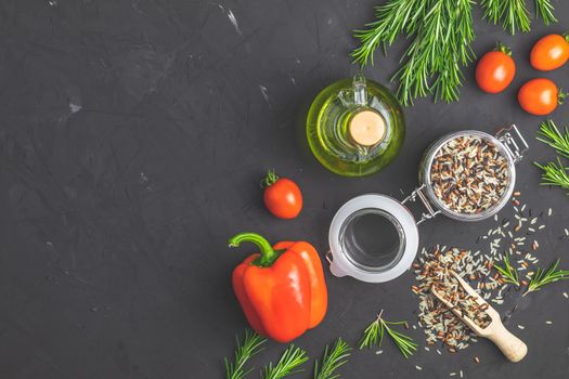 Black, purple, and white rice (Oryza sativa) mix in glass jar on black stone concrete textured surface background. Raw pepper, tomatoes, olive oil and rosemary bunch. Top view with copy space for your text.