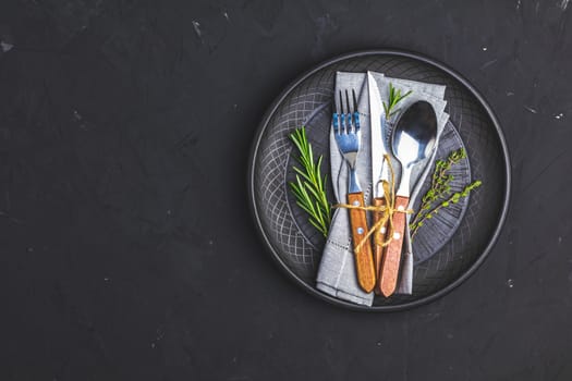 Rustic vintage set of cutlery knife, fork and spoon in black ceramic plate. Black stone concrete surface background. Top view, copy space.