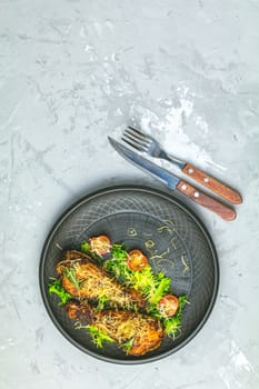 Baked chicken drumstick in a black ceramic plate with tomatoes and rosemary, light gray stone concrete surface, top view, copy space.