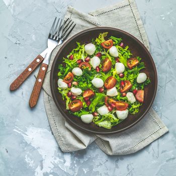 Fresh Cherry Tomato, Mozzarella salad with green lettuce mix served on a brown ceramic plate, healthy food, light gray stone concrete surface, top view, copy space.