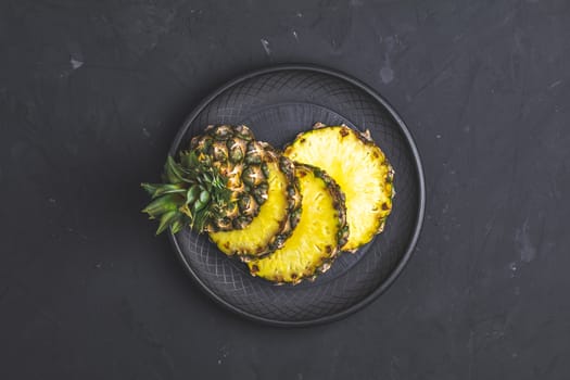 Sliced pineapple in ceramic plate on black stone concrete textured surface background. Top view with copy space for your text.