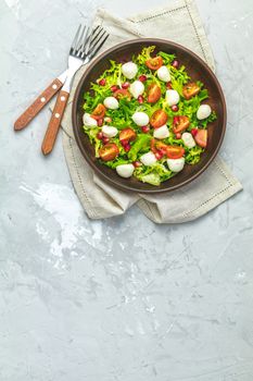 Fresh Cherry Tomato, Mozzarella salad with green lettuce mix served on a brown ceramic plate, healthy food, light gray stone concrete surface, top view, copy space.