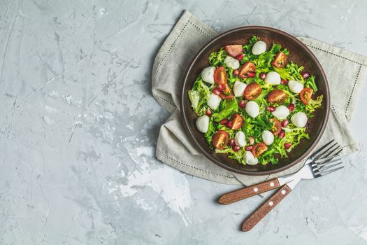 Fresh Cherry Tomato, Mozzarella salad with green lettuce mix served on a brown ceramic plate, healthy food, light gray stone concrete surface, top view, copy space.