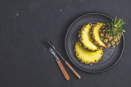 Sliced pineapple in ceramic plate and set of cutlery knife, fork  on black stone concrete textured surface background. Top view with copy space for your text.