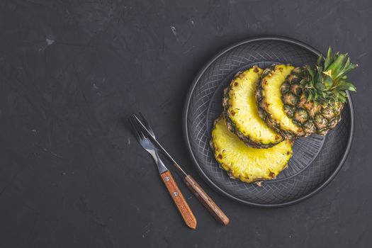 Sliced pineapple in ceramic plate and set of cutlery knife, fork  on black stone concrete textured surface background. Top view with copy space for your text.