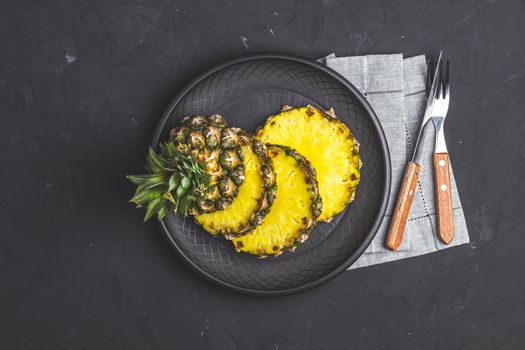Sliced pineapple in ceramic plate and set of cutlery knife, fork  on black stone concrete textured surface background. Top view with copy space for your text.