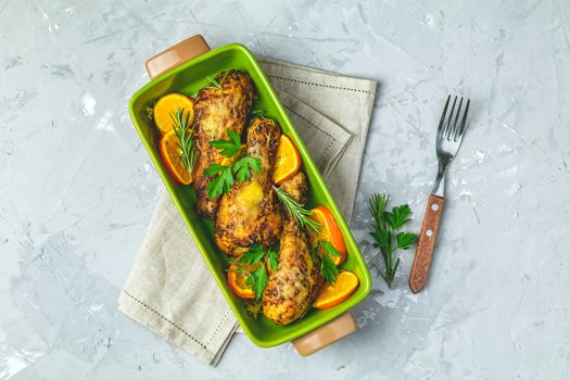 Baked chicken drumstick in a green dish with orange and rosemary, light gray stone concrete surface, top view, copy space.