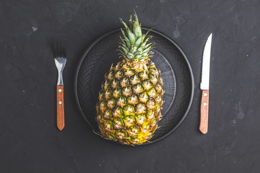 Pineapple in ceramic plate and set of cutlery knife, fork  on black stone concrete textured surface background. Top view with copy space for your text.