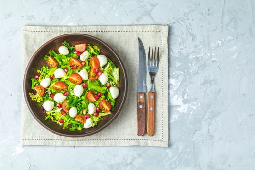 Fresh Cherry Tomato, Mozzarella salad with green lettuce mix served on a brown ceramic plate, healthy food, light gray stone concrete surface, top view, copy space.