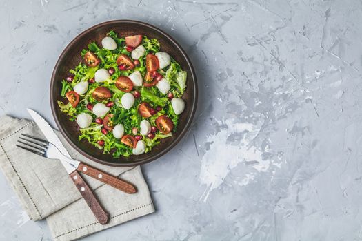 Fresh Cherry Tomato, Mozzarella salad with green lettuce mix served on a brown ceramic plate, healthy food, light gray stone concrete surface, top view, copy space.