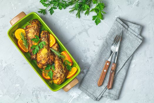 Baked chicken drumstick in a green dish with orange and rosemary, light gray stone concrete surface, top view, copy space.