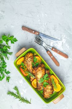 Baked chicken drumstick in a green dish with orange and rosemary, light gray stone concrete surface, top view, copy space.