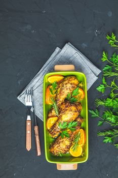 Baked chicken drumstick in a green dish with orange and rosemary, black stone concrete surface, top view, copy space.