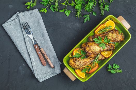 Baked chicken drumstick in a green dish with orange and rosemary, black stone concrete surface, top view, copy space.