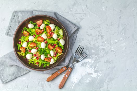 Fresh Cherry Tomato, Mozzarella salad with green lettuce mix served on a brown ceramic plate, healthy food, light gray stone concrete surface, top view, copy space.