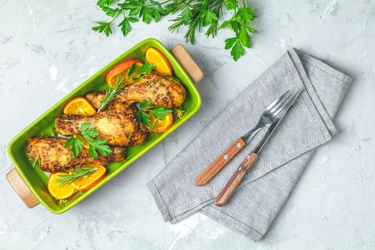 Baked chicken drumstick in a green dish with orange and rosemary, light gray stone concrete surface, top view, copy space.