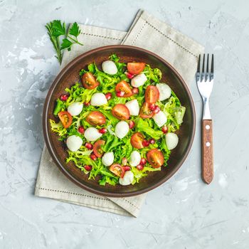 Fresh Cherry Tomato, Mozzarella salad with green lettuce mix served on a brown ceramic plate, healthy food, light gray stone concrete surface, top view, copy space.